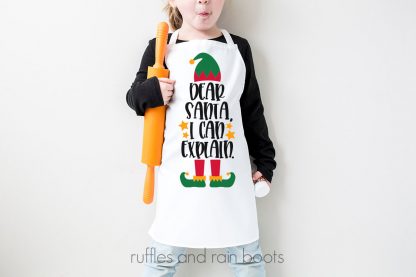 horizontal image of child in apron against white wall with Christmas SVG that says Dear Santa I can explain