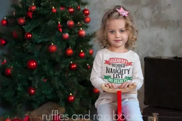 horizontal image of Christmas tree background with little girl wearing a sweatshirt with the design legendary member of the naughty list svg with banners