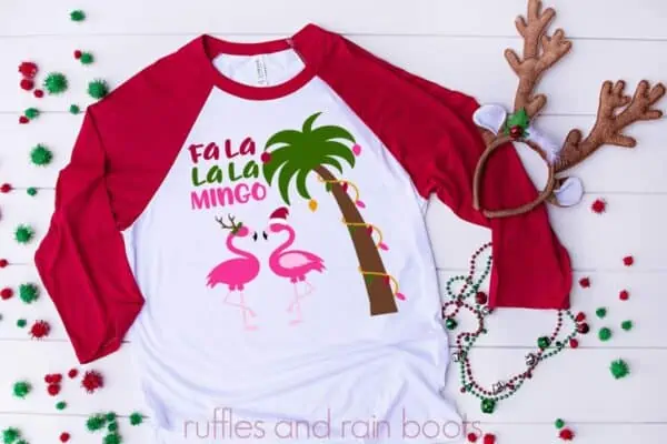 horizontal image of white shirt with red raglan sleeves on holiday background with flamingo Christmas svg featuring text which reads fa la la la mingo, two flamingoes in holiday accessories, and a palm tree in Christmas lights