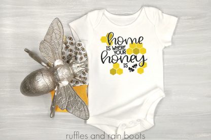 horizontal image of white onesie on wood background with bee and home is where your honey is svg in vinyl