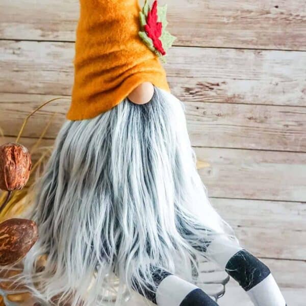 Close up image of a fall shelf sitter gnome pattern made with felt hat, Mongolian fur in frosted gray, and long black and white striped legs in front of a wood background.