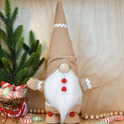 Adorable gingerbread gnome with white beard, shoes, and rick rack trim in front of a holiday background with candy, farmhouse beads, and tree.