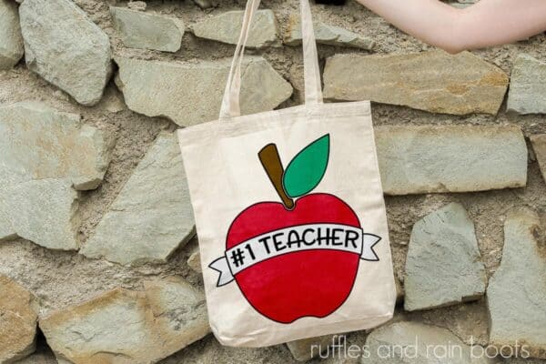horizontal image of number 1 teacher svg in white banner on red apple placed in sublimation on a tote bag being held in front of rock wall
