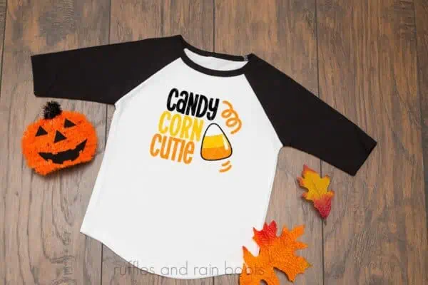 horizontal image of wood background with halloween svg candy corn cutie in black orange and yellow on black raglan sleeve shirt