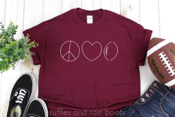horizontal of maroon t shirt with elegant minimalist peace love football svg in white vinyl on a white wood background with football jeans and tennis black tennis shoes
