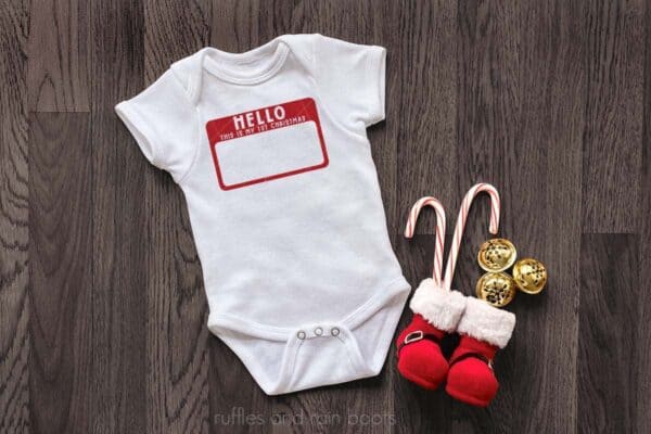 horizontal image of white bodysuit with Hello this is my first Christmas on the iconic hello sticker in red on a dark wood background with santa boots filled with candy canes and gold sleigh bells