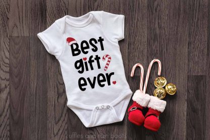 horizontal of a white baby bodysuit with the best gift ever SVG in black and the Santa Hat SVG, candy cane, and heart SVG in red shown on a dark wood background with holiday Santa boots and bells