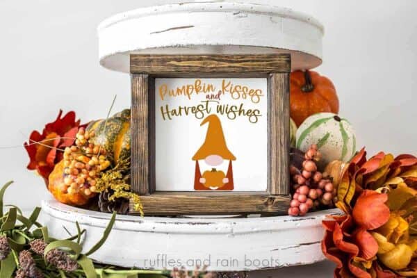 horizontal image of fall gnome svg bundle with fall gnome holding a pumpkin on a white and wood sign placed on a fall themed tiered tray background with text which reads pumpkin kisses and harvest wishes