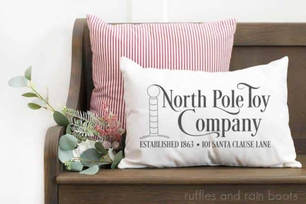 horizontal image of white pillow with north pole toy company in HTV vinyl cut with Cricut machine on wood bench in front of holiday greenery and red and white ticking stripe pillow