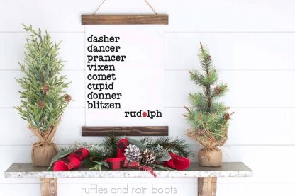 horizontal image of scroll Christmas sign with Santa reindeer names in black and red hanging on white shiplap wood wall over a holiday mantle decorated with buffalo check, holiday ornaments, and Christmas trees