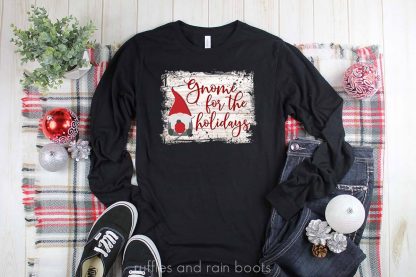 horizontal image of gnome for the holidays sublimation pressed on a black t shirt over white glitter heat transfer vinyl on a Christmas background on white wood with blanket and ornaments