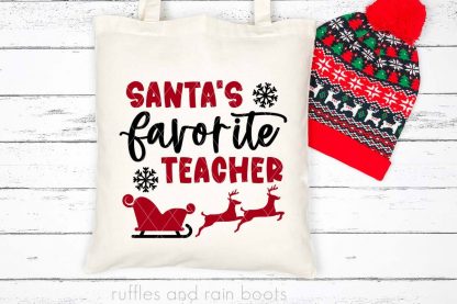horizontal image of a close up a canvas tote bag with a red and black Santa's favorite teacher SVG sleigh and reindeer in vinyl on a wood background with holiday hat
