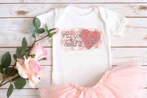 close up horizontal image of a wood background with pink rose pink tutu and white baby bodysuit with the all you need is love sublimation design in plaid and glitter