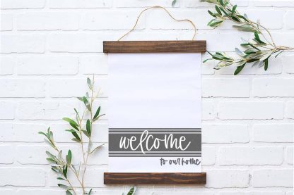 horizontal image showing a white canvas hung between two dark wood slats which reads welcome to our home in a dark gray vinyl on a white brick background with pale green leaves