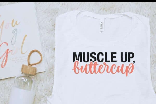 horizontal image of a white muscle up buttercup svg in black and pink on white muscle tank top for workouts sitting on a cream flokati rug next to a white and glass water bottle