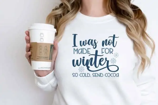 horizontal image of woman in front of white wall wearing a white sweatshirt with blue vinyl svg of I was not made for winter cut file with snowflakes and so cold send cocoa