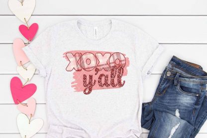 horizontal image of light gray shirt on white wood Valentine background with plaid and glitter xoxo y'all sublimation print in pinks and reds