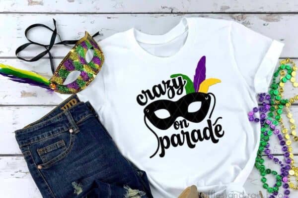 white t shirt with black purple green and yellow vinyl in the shape of a mask with feathers on one side with text crazy on parade on wood background with mardi gras beads and a sequined mask.