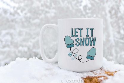 horizontal image of a white coffee mug with let it snow in shades of teal and black vinyl with mittens and snowflakes in the snow against back drop of snow covered trees.