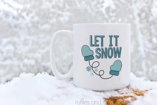 horizontal image of a white coffee mug with let it snow in shades of teal and black vinyl with mittens and snowflakes in the snow against back drop of snow covered trees.