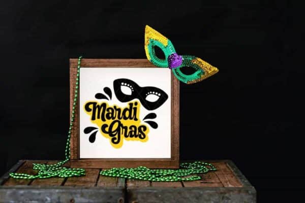 close up horizontal image of a free mardi gras offset svg in yellow and black vinyl placed on a white sign on top of a wood trunk with beads and a sequin mask and black background.