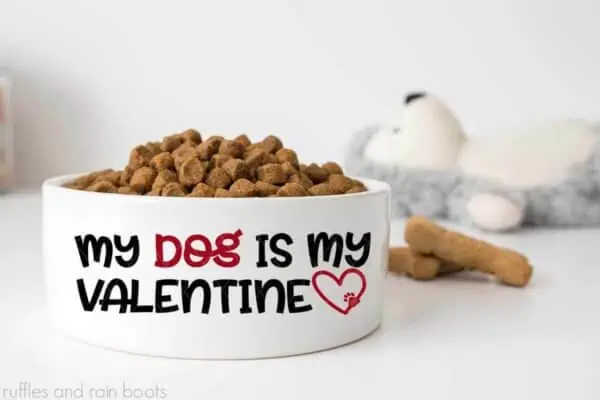 close up horizontal image of a dog bowl with kibble a treat and a stuffed animal with text which reads my dog is my valentine svg from ruffles and rain boots