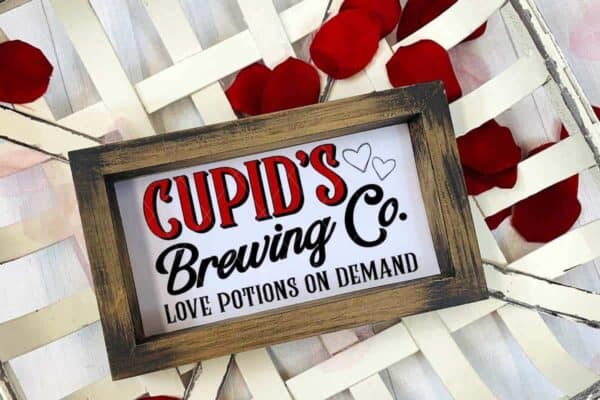 Small wooden sign in a tobacco basket with rose petals and red and black vinyl which reads Cupids Brewing Company Valentine SVG with hearts and love potions SVG from ruffles and rain boots.