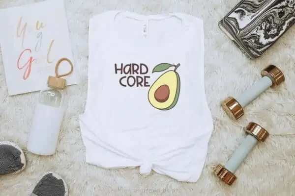 Horizontal image showing hard core avocado svg on white muscle shirt on workout background with water bottle, weights, and tennis shoes.
