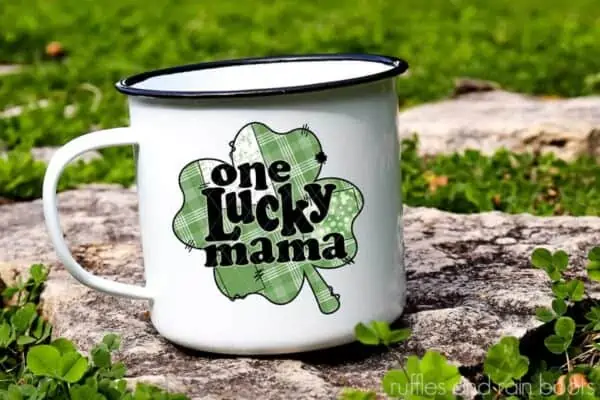 Horizontal image of a one lucky mama shamrock sublimation transferred onto a white enamel camping mug with black rim sitting on rock in field of clover.