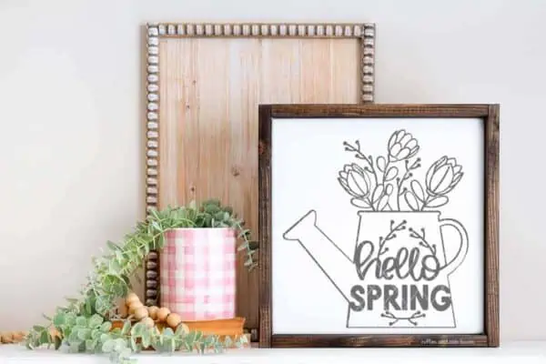 Close up horizontal image of a gray hello spring watering can svg in front of a wood plank and garden pot stacked on books filled with succulent.