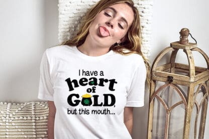 Horizontal close up image of a blonde woman sticking her tongue out with eyes closed while wearing a white t shirt with the vinyl saying I have a heart of gold but this mouth in black, gold, and green vinyl.