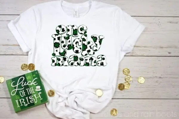 Horizontal image of white t shirt with green and black one lucky mama sublimation print with leopard print shamrocks on a wood background with gold coins and a St Patricks day sign.
