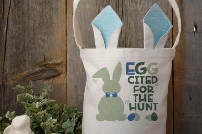 Horizontal image of a bunny ear Easter basket on a dark wood background with EGGcited for the hunt SVG with bunny and eggs in green and blue vinyl.