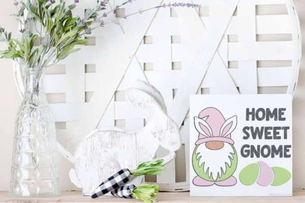 Horizontal image of a holiday display with farmhouse style basket, wood bunny, and buffalo plaid carrots and Home Sweet Gnome Easter SVG on white wood sign in pink, green, and gray.