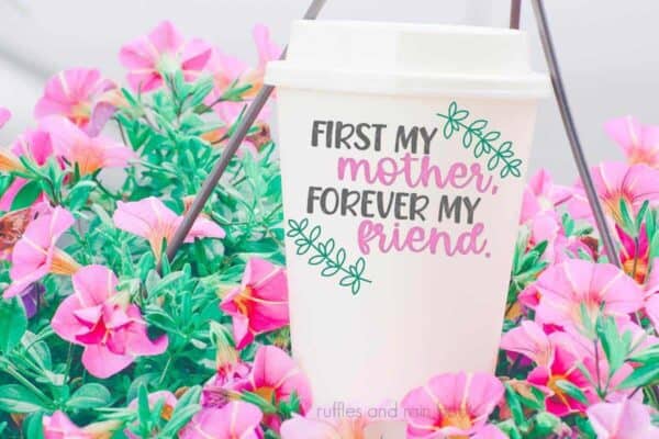 Horizontal close up image of a white travel cup with gray, pink, and green first my mother, forever my friend svg with green laurels sitting in a hanging pot full of pink flowers.