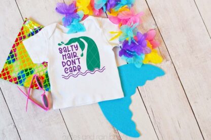 Horizontal image showing a mermaid tail and lei of flowers on wood background with child's white t-shirt with salty hair don't care mermaid SVG free bundle element and text which reads ruffles and rain boots.