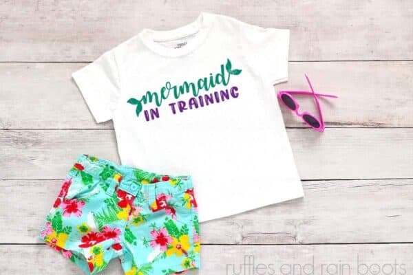 Horizontal image showing floral shorts and sunglasses on white wood background with white girl's t-shirt with mermaid in training SVG in teal and purple as part of the mermaid SVG free bundle from ruffles and rain boots.
