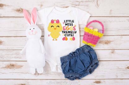 Horizontal image of a white body suit with yellow pink orange and black heat transfer vinyl with Eggs tremely cute Easter design with basket bunny and jean shorts on white wood background.