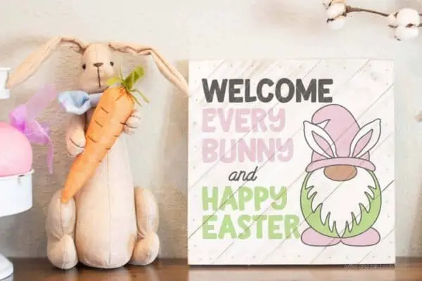 Horizontal image of an Easter Welcome sign SVG with bunny gnome cut file on a white wood sign with a rustic bunny plush holding a carrot.