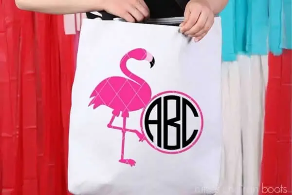 Horizontal image of a girl wearing a canvas tote bag with a pink flamingo monogram and initials in black in front of background of party streamers in red, white, and teal.