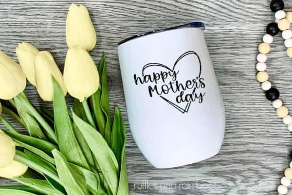 Horizontal image of a white tumbler with black vinyl of happy mother's day SVG lying on a wood background next to yellow tulips and a set of neutral farmhouse beads.