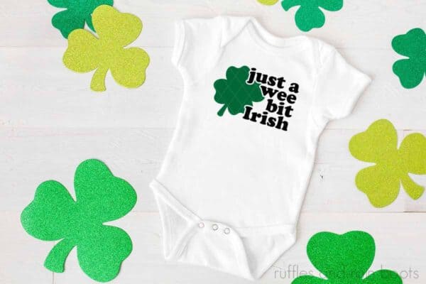 Horizontal image of a white baby body suit with a green shamrock and black lettering which reads just a wee bit Irish.
