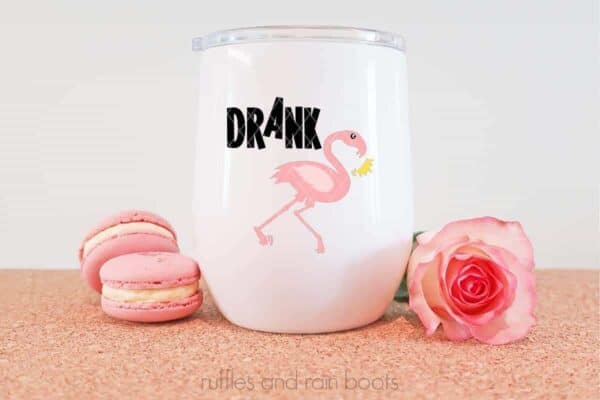 Horizontal image of a falling flamingo losing her crown with the word Drank in black vinyl on a white tumbler and a cork background.