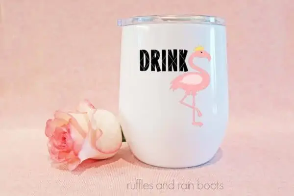 Horizontal image of a pink flamingo with a crown SVG with DRINK in black permanent vinyl on a white tumbler on pink background with pink rose.