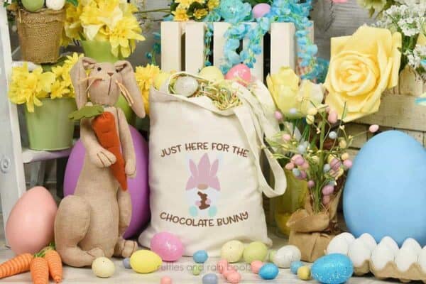 Horizontal image showing a floral and egg background with a stuffed bunny next to a tote bag with an Easter gnome with the saying just here for the chocolate bunny SVG in pastels.