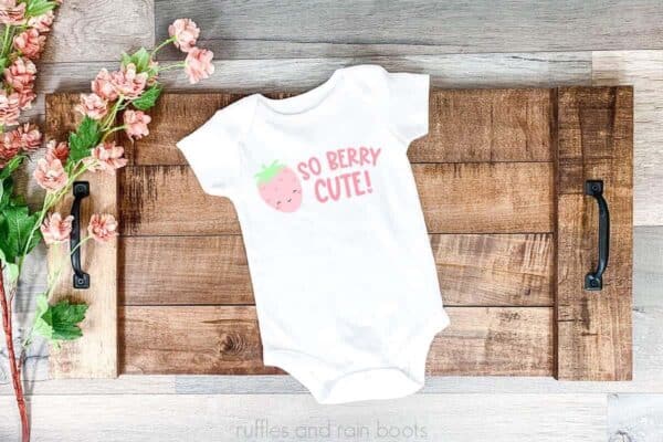 Horizontal image showing flowers and a wood tray with a white bodysuit decorated in vinyl with a cute Kawaii strawberry and the saying so berry cute in pink.