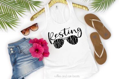 Horizontal image showing palm fronds, flip flops, and jean shorts on white background with white tank with black vinyl sunglasses with Resting Beach Face SVG cut out.