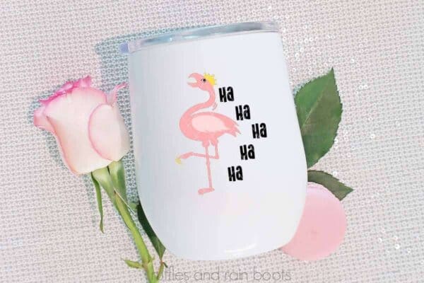 Horizontal image of a white tumbler with a laughing flamingo SVG with a crown on a white sparkly background with a pink rose and macaron.