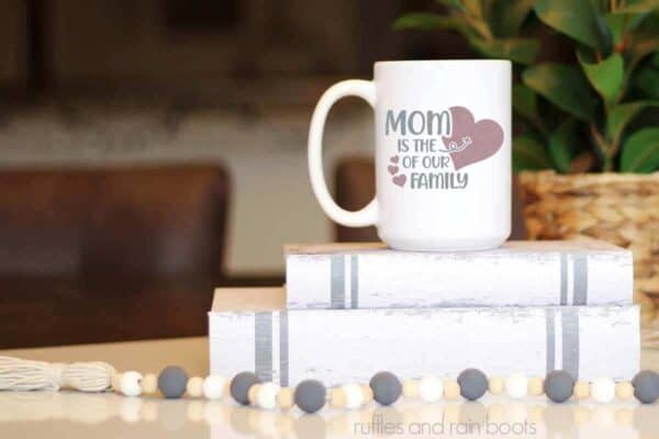 Horizontal image showing a close up of a gray and mauve mother's day ceramic mug with mom is the heart of our family SVG in vinyl on top of book stack and farmhouse beads.