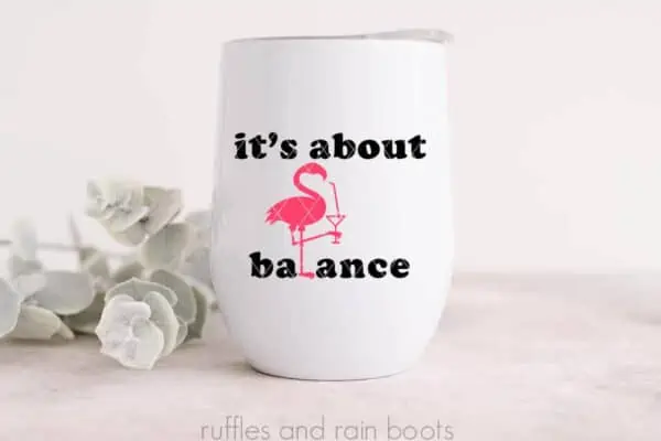 Horizontal image of a pink vinyl flamingo holding a cocktail with the words it's about balance in black vinyl on a white stemless wine tumbler in front of some greenery on a white background.